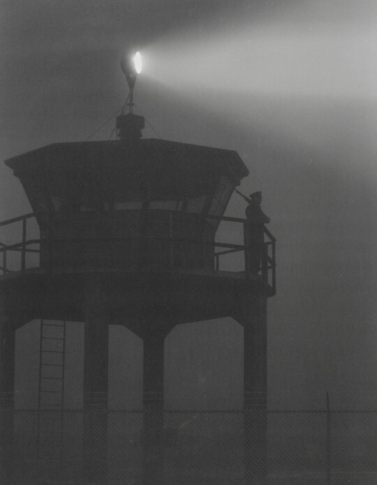 A black-and-white photograph of a person standing in silhouette atop a tower with a bright spotlight shining from the top.
