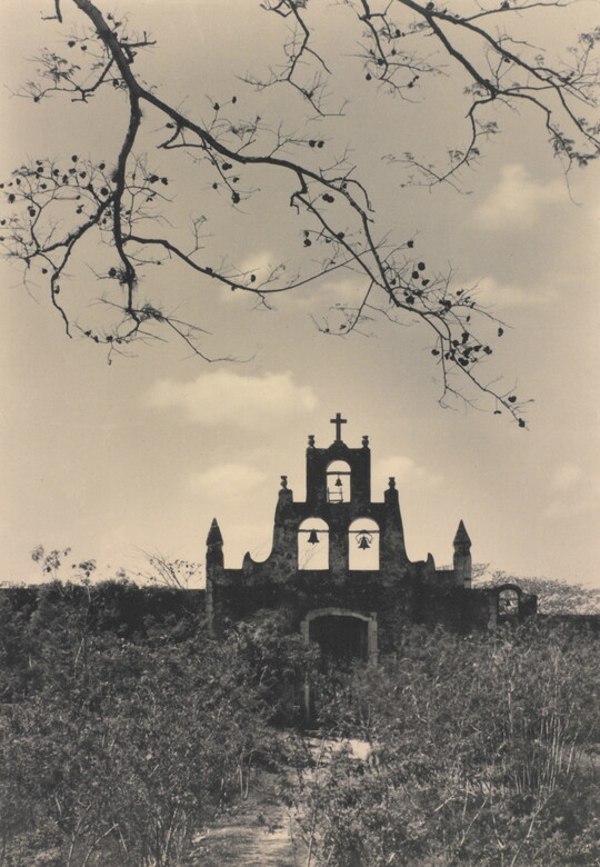 A black-and-white photograph of the facade and terraced bell tower, topped with a cross, of an adobe church in a field of tall grass and weeds.