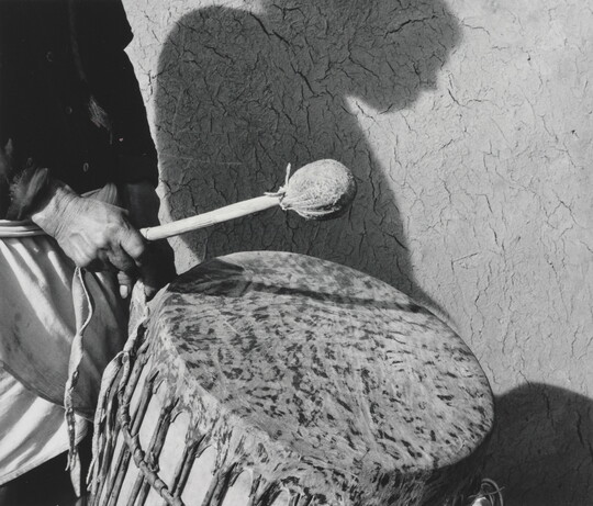 A black-and-white photograph of a person holding a drumstick over a traditional drum and the person's shadow in profile on the wall in the background.