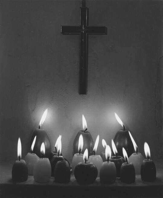 A black-and-white photograph of three rows of lit votive candles in front of a cross hanging on a wall.