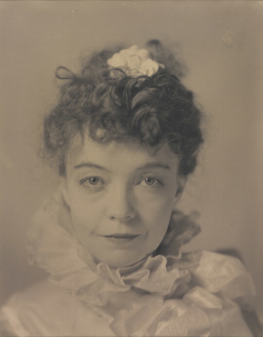 A black-and-white portrait photograph of a White woman with curly hair pulled on top of her head, wearing a top with a ruffled collar.