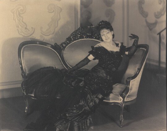 A black-and-white photograph of a White woman in a long, formal gown and elbow-length gloves posing on a sofa.