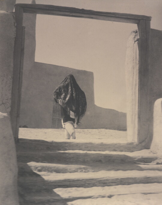 A black-and-white photograph of a person wearing a long shawl over their head and torso while walking through the wood gate of an adobe building.
