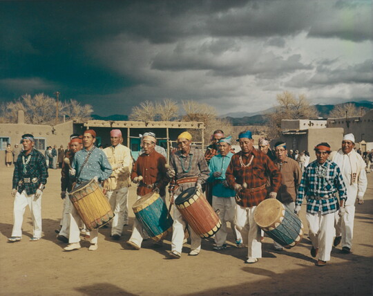 A color photograph of Indigenous people walking, four of them carrying tall drums.