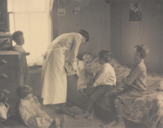 A black-and-white photograph of a nurse bending over a person in a bed, two children sitting on the bed, and two children behind the nurse.