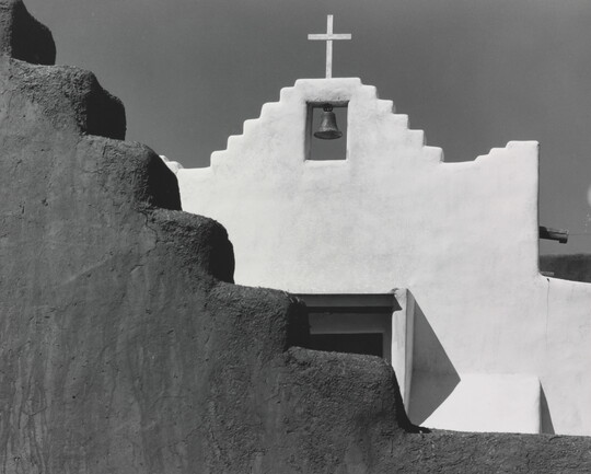 A black-and-white photograph of a terraced adobe bell tower with a cross on top, partially hidden by a terraced adobe wall.