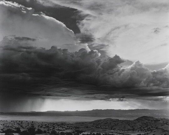 A black-and-white photograph of torrential rain and heavy storm clouds passing into a brightly lit plain.