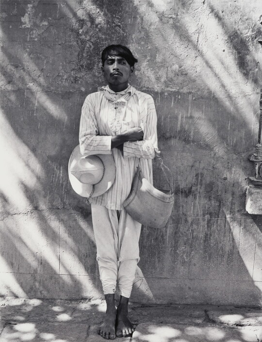 A black-and-white photograph of a dark-skinned man in light-colored shirt and pants, holding a hat under one crossed arm and a bag on the other, leaning against a wall.