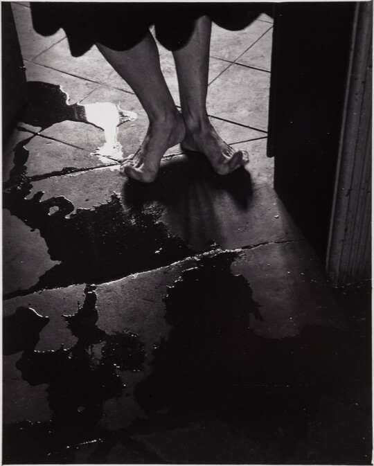 A black-and-white photograph of a person's bare feet, toes stretched up off the tile floor to avoid puddles.