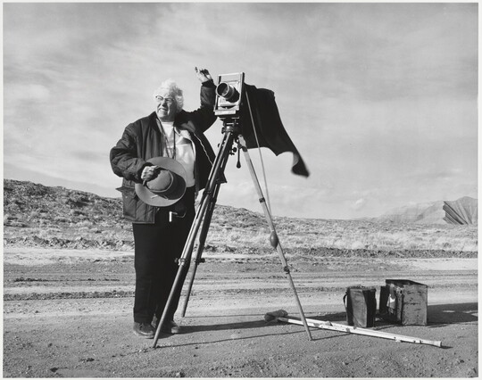 A black-and-white photograph of an older White woman holding a cowboy hat and standing next to a large format camera on a dirt road.