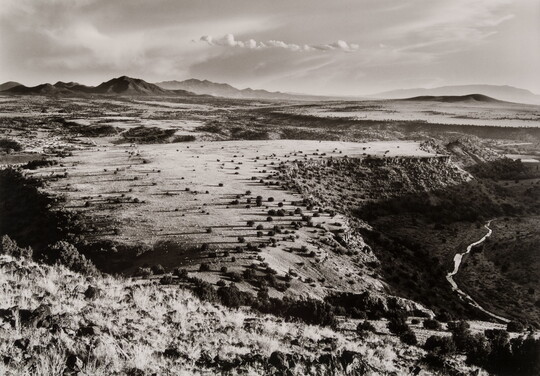 A black-and-white photograph of the top of a mesa with a winding river below and mountains in the distance.