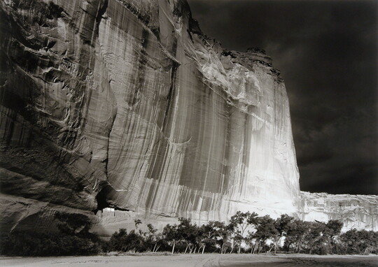 A black-and-white photograph of a sheer cliff face at night with a small ruined structure near the bottom, and trees on the plain in front of the cliff.