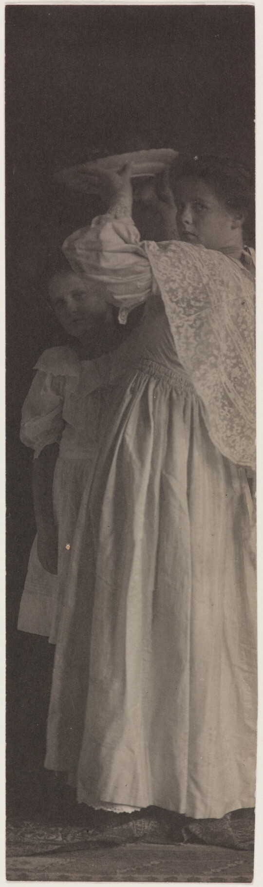 A very vertically-oriented black-and-white photograph of two White girls in formal dresses, one holding a plate above her head.