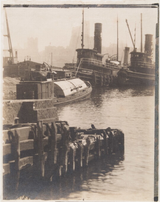 A black-and-white photograph of a narrow wharf with boats alongside and tall buildings in the distance.