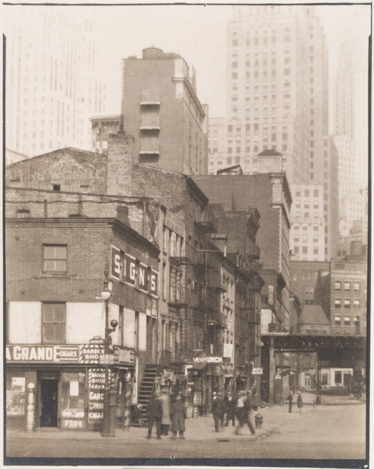 A black-and-white photograph of people on a street corner near three- and four-story buildings with skyscrapers in the background.
