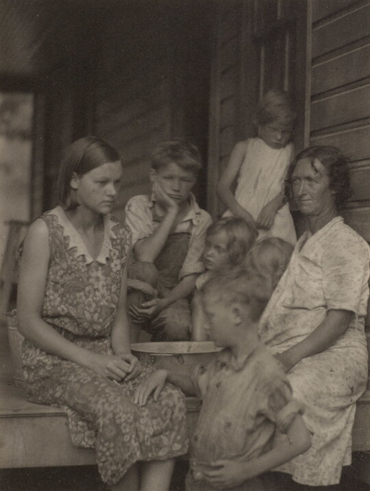 A black-and-white photograph of two White women and several children on the porch of a clapboard house.