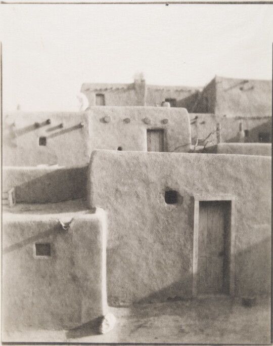 A black-and-white photograph of a multi-level adobe pueblo with wood doors in some of the walls.