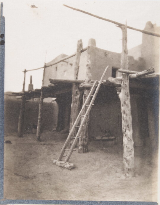 A black-and-white photograph of a multi-level adobe pueblo with a wood ladder and wood framing supporting covered porches.