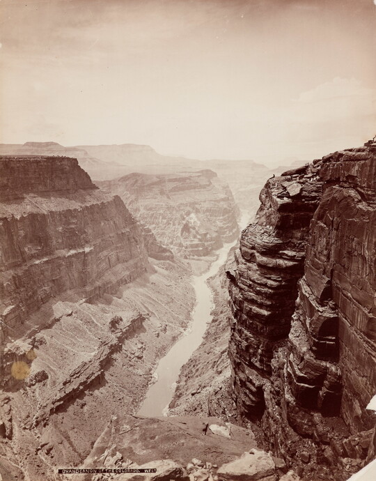 A sepia-toned photograph of a river at the bottom of a steep canyon with sheer cliffs on both sides.