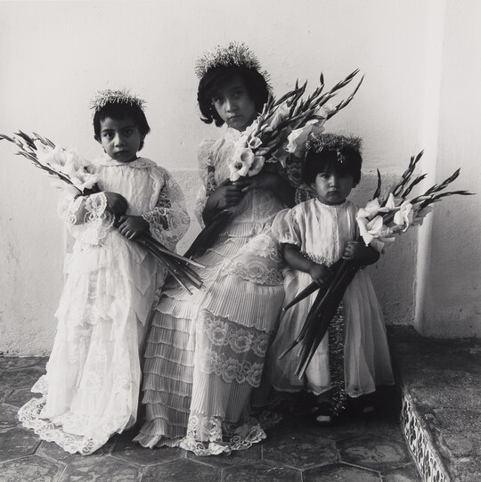 A black-and-white photograph of three young medium-skinned girls wearing floral tiaras, light-colored dresses, and holding bouquets of lilies.