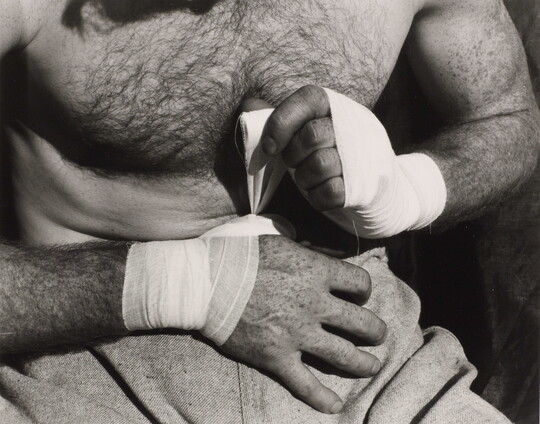 A black-and-white, close-up photograph of a light-skinned, shirtless boxer wrapping his hands with tape.