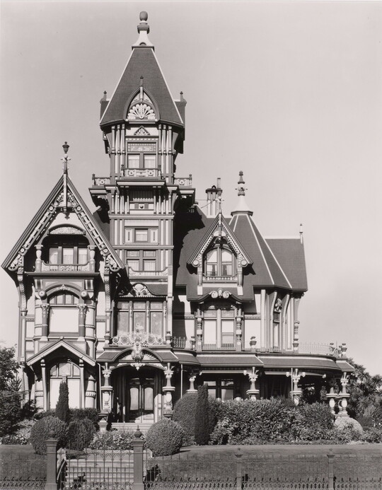 A black-and-white photograph of a large, multi-story, elaborate Victorian house with turrets and a wrap-around porch on a sunny day.