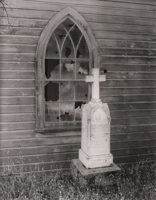 A black-and-white photograph of a tall, white tombstone with a cross on top next to a wood building with a broken lancet-shaped window.