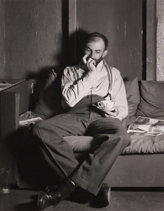 A black-and-white portrait photograph of a bearded White man seated on a sofa with one hand up to his mouth, the other holding a teacup over his lap.