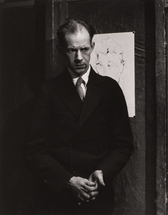 A black-and-white portrait photograph of a balding White man wearing a coat and tie holding his hands clasped in front of him.
