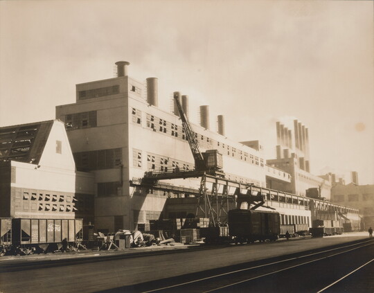 A black-and-white photograph of a multi-story industrial building with multiple smokestacks on top and tracks for a streetcar in front.