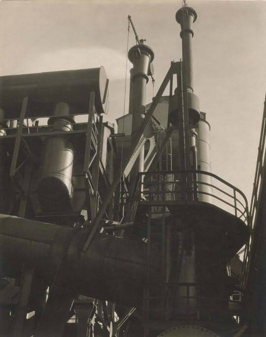 A black-and-white photograph of the exterior of a factory taken from the ground looking up toward the top of its stacks.