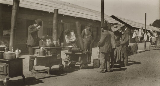 A black-and-white photograph of a group of people, mostly men in bowler hats, standing around outdoor stoves lined up in front of a row of low, long buildings.
