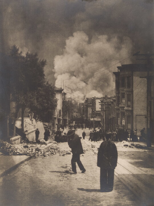 A black-and-white photograph of people standing in a street near piles of rubble and billowing smoke in the distance.