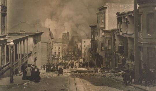 A black and white photograph of people standing in a street filled with rubble from ruined buildings on either side as smoke billows over buildings at the end of the street.