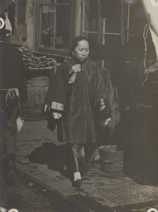 A black-and-white photograph of an woman with Asian features wearing a billowing tunic over pants walking down a commercial street.
