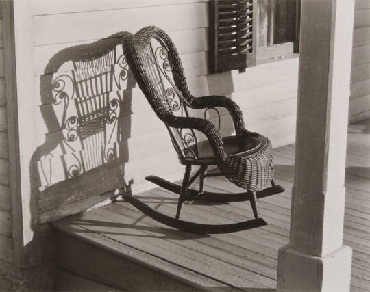A black-and-white photograph of a decorative wicker rocking chair on a porch that casts a stark shadow on the clapboard behind it.