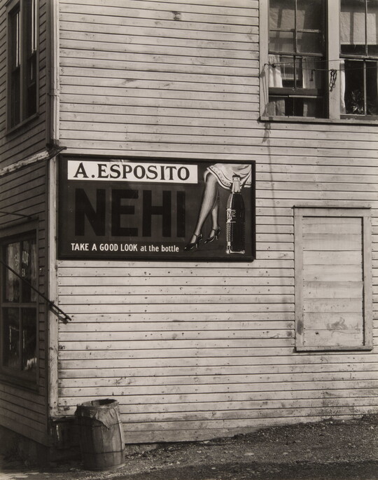A black-and-white-photograph of the side of an old, clapboard building with a large sign advertising a soft drink.