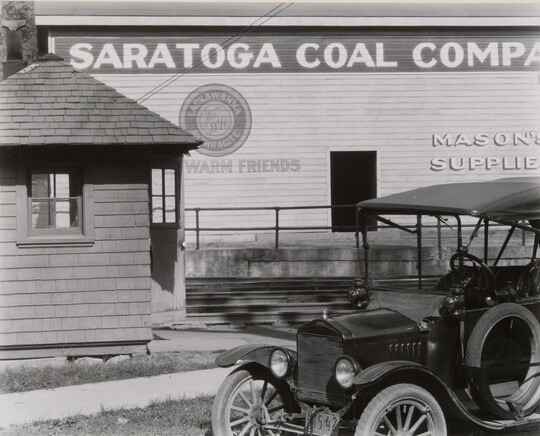 A black-and-white-photograph of a 1920's-style automobile parked in front what looks like a warehouse that is next to railroad tracks.