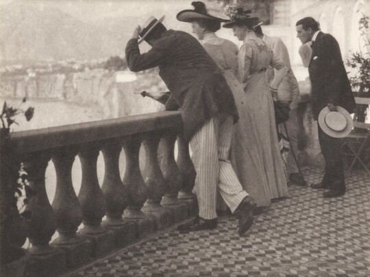 A black-and-white photograph of a group of well-dressed people looking over a balcony with a rugged coastline in the distance.
