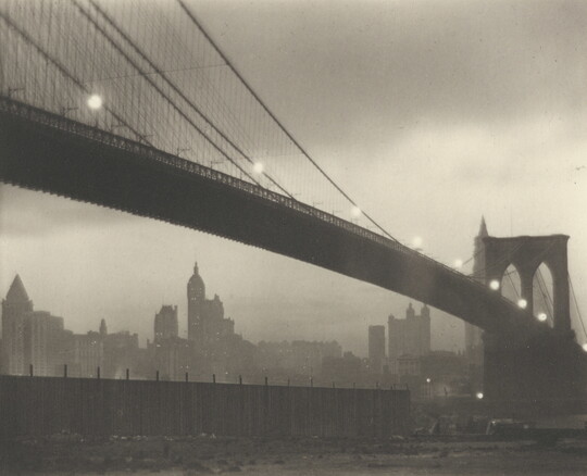 A sepia-toned photograph of a long bridge with a foggy cityscape in the background at nighttime.