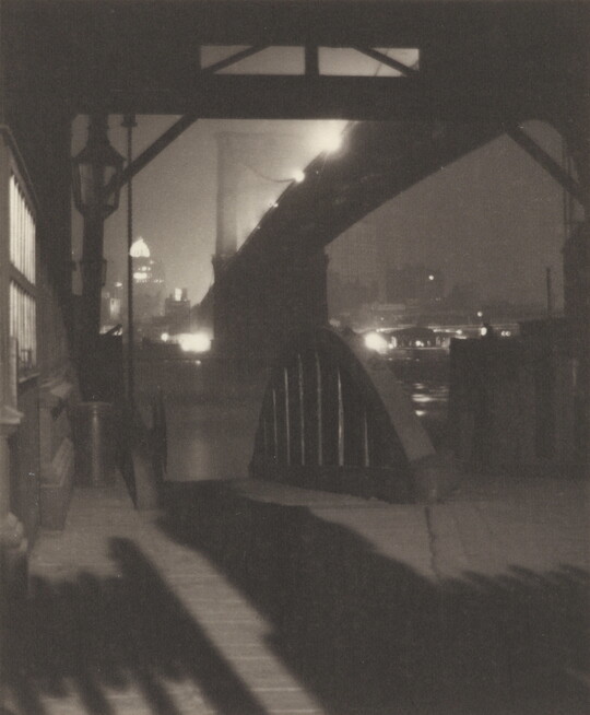 A black-and-white photograph of a dock underneath a large suspension bridge illuminated by street lights.