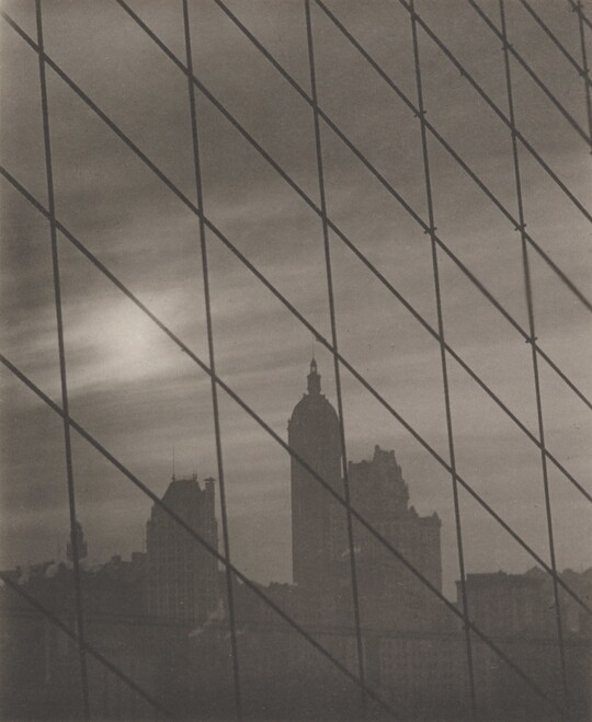 A black-and-white photograph of a grid of cables framing a cityscape with tall buildings in the background.