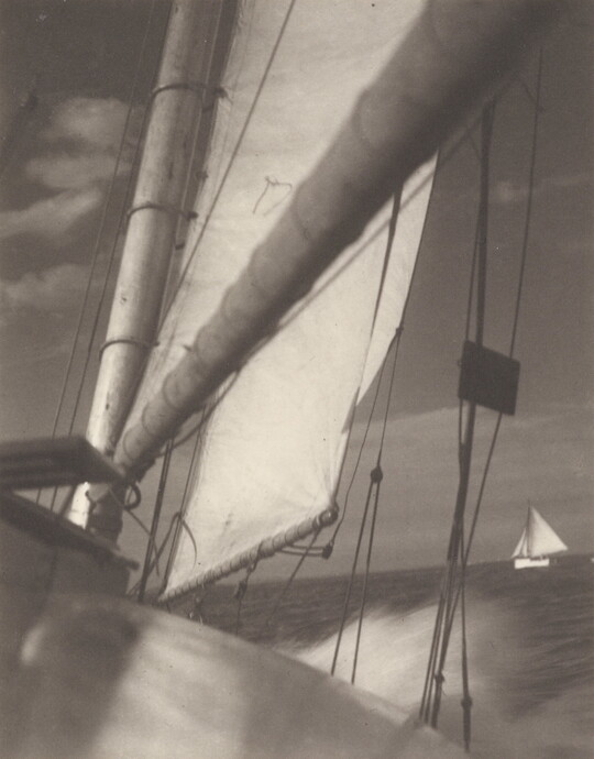A black-and-white photograph looking down the boom of sailboat and sail at another sailboat on the sea in the distance.