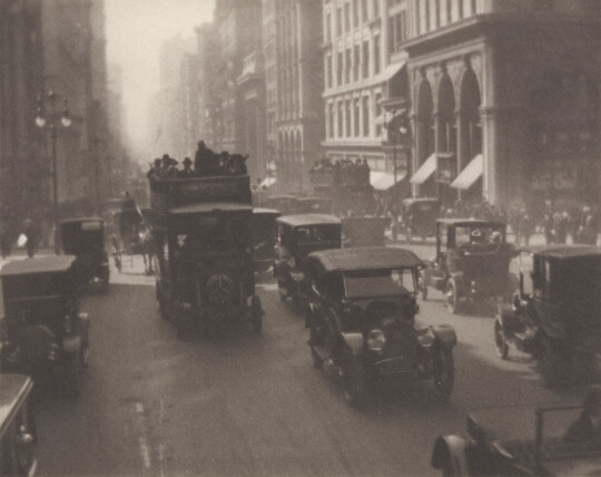 A black-and-white photograph of early 20th-century cars, busses, and wagons pulled by horses on a busy city street.