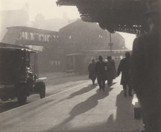A black-and-white photograph of people walking along the sidewalk of a city street as the sunlight casts long shadows.
