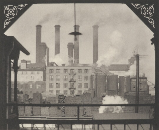 A black-and-white photograph of a cluster of industrial buildings with tall billowing smoke stacks, as viewed from a balcony.