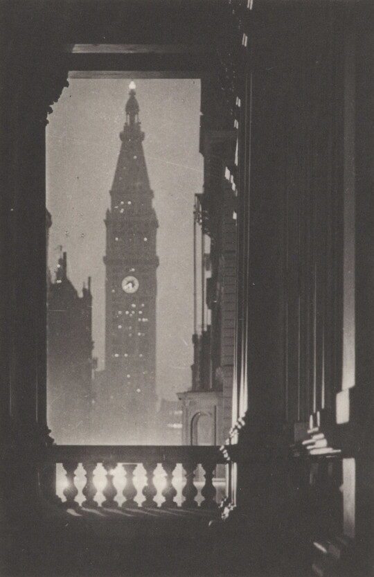 A black-and-white photograph of a tall, glimmering skyscraper at night as seen through an interior door, framed woodwork and a banister.
