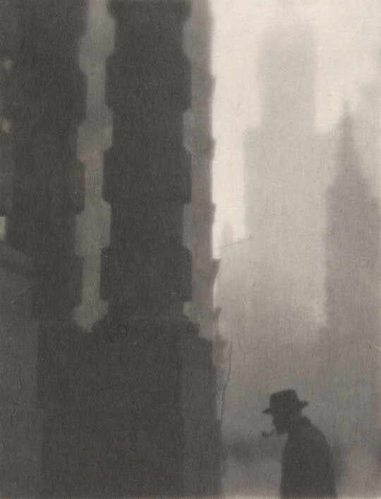 A black-and-white photograph of the silhouette of a man smoking a pipe next to towering columns and buildings.