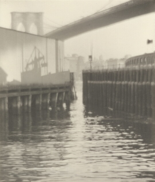A black-and-white photograph of the shadow of a ship on top of a pier in a busy harbor.