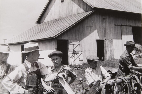 A black-and-white photograph of White men and boys dressed in farming clothes arranging items on a hitching post outside of a barn.